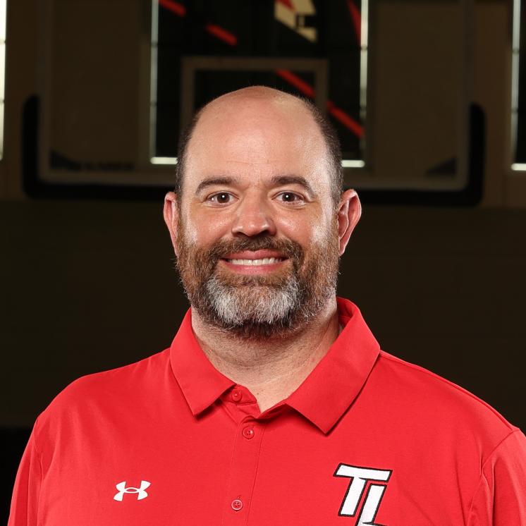 A man stood indoors in athletic clothing with a basketball goal behind him