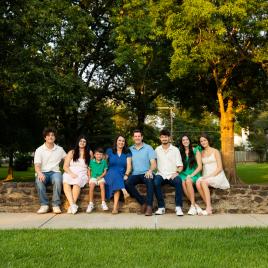 Picture of a family sitting on a stone retaining wall with green trees and green grass around them. 
