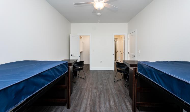 A minimalist dorm room with two beds, desks, and chairs, featuring white walls, dark floors, and a ceiling fan
