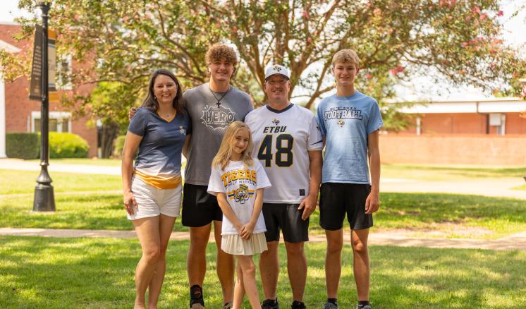 A family stood together outside on green grass underneath a tree with outdoor attire
