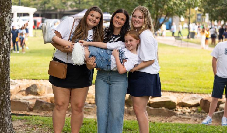 3 women stood outside on green grass holding a child in a plank pose