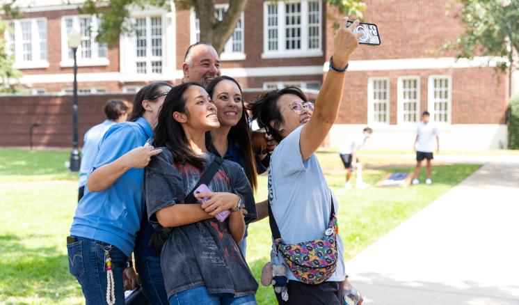 A group of people stood outside together arms outstretched holding a cell phone for a "selfie"
