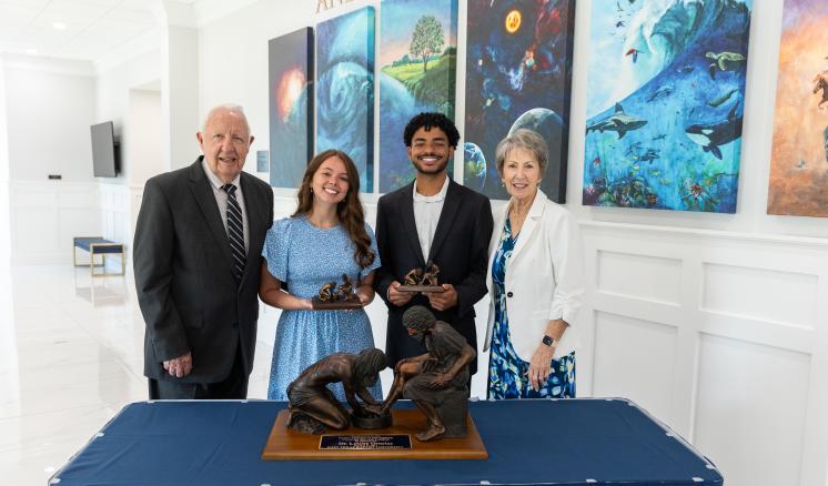 Bob and Gayle Riley stand on either side of a male and female student holding awards behind a table inside a building with white walls.