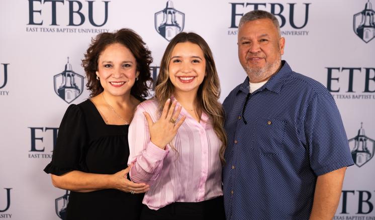 3 people standing in front of an ETBU backdrop with the center persons hand extended showing a ring
