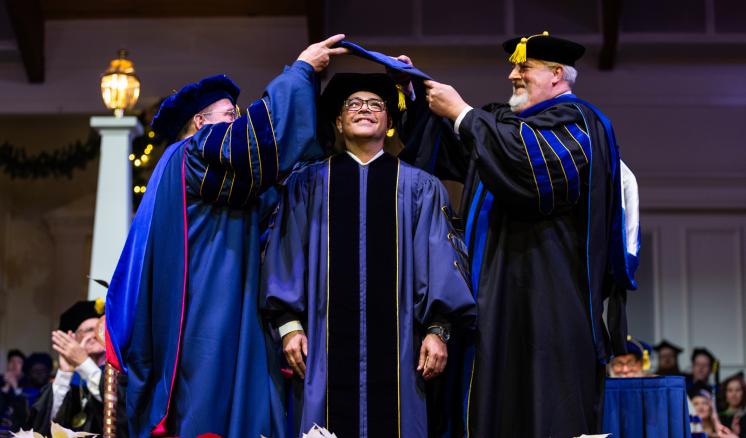 Three men in graduation regalia with the left and right men hooding a doctoral student in the middle.