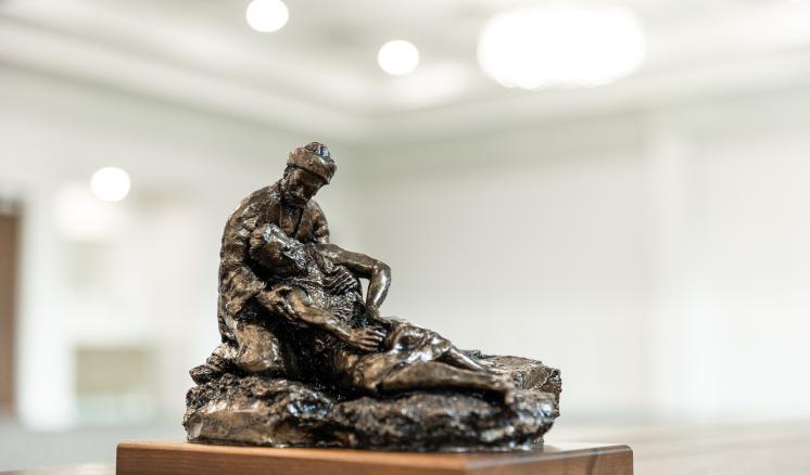 Bronze sculpture of the Good Samaritan kneeling and cradling an injured man, displayed on a wooden base in a softly lit indoor space.