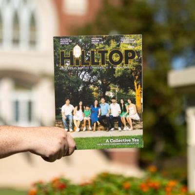 A hand holding the Hilltop Magazine 2025 edition in front of a red brick building and green trees on a sunny day.