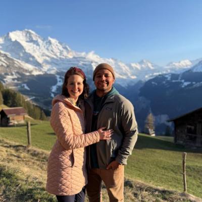 2 people standing in a grass field with snow capped mountains behind them
