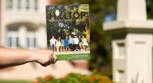 A hand holding the Hilltop Magazine 2025 in front of a brick building with plants blurred in the background.