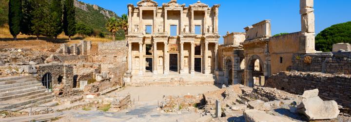 The ancient Library of Celsus in Ephesus, Turkey, with its grand two-story marble façade, columns, and arched entryways surrounded by ruins and stone walkways under a clear blue sky