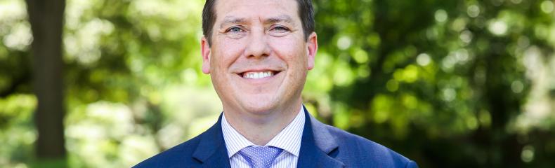 A man in formal clothing stood outside in front of trees on a walking path