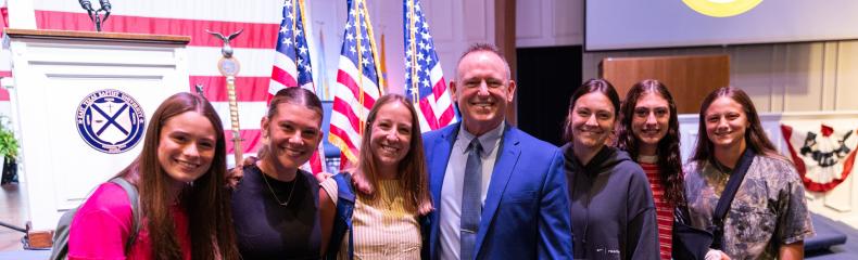 A group of people standing together smiling with the backdrop of American Flags on a stage