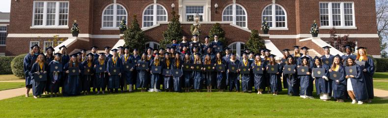 Group photo of graduates in navy regalia in front of Marshall Hall on a sunny day.