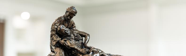 Bronze sculpture of the Good Samaritan kneeling and cradling an injured man, displayed on a wooden base in a softly lit indoor space.